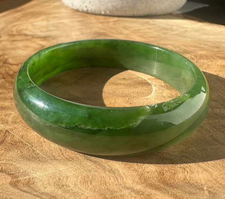 A round green jadeite bangle bracelet with natural marbling, resting on a wooden surface under direct sunlight.