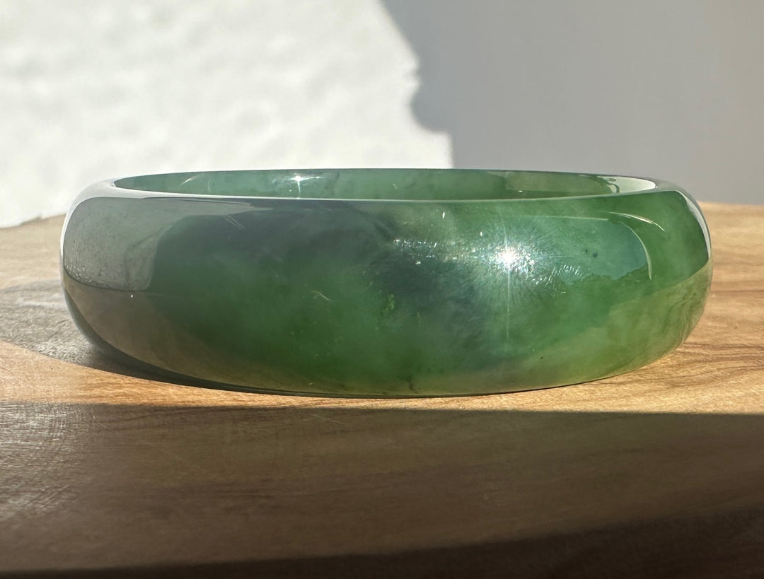 A polished green jade bangle bracelet resting on a wooden surface with a white textured background.