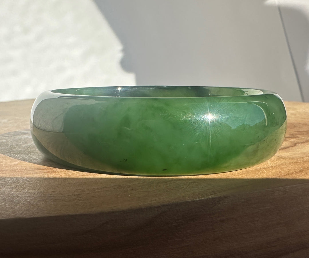 A polished green jade bangle bracelet resting on a wooden surface with natural sunlight casting soft shadows.