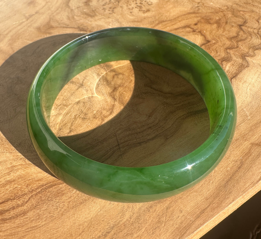 A round green jade bangle bracelet with a glossy finish, resting on a natural wood surface under direct sunlight.