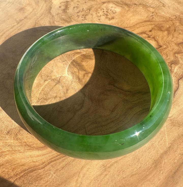 A round green jade bangle bracelet displayed on a natural wood surface, showing a glossy finish and natural color variations.