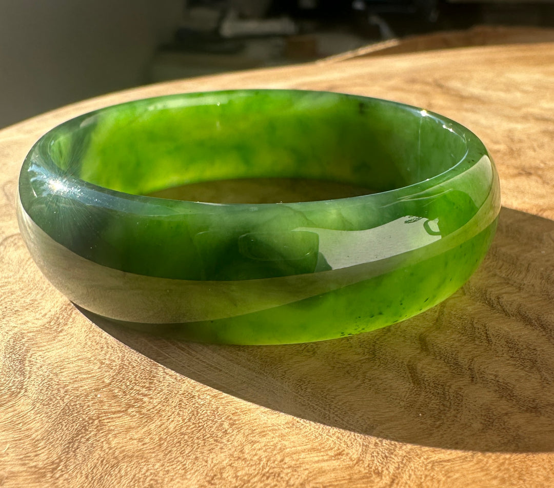 A polished green jade bangle bracelet resting on a light brown wooden surface under direct sunlight.