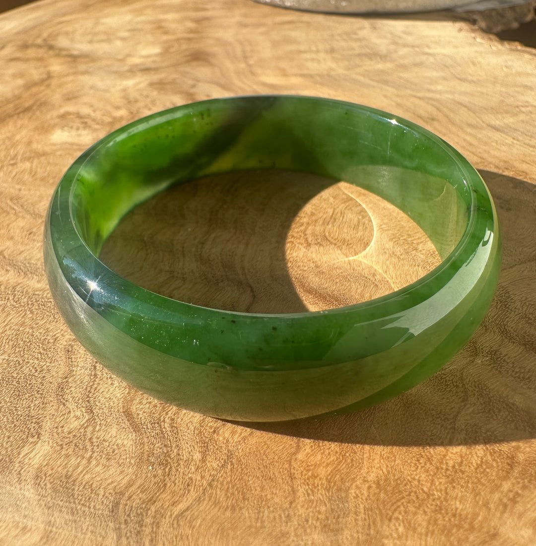 A round green jade bangle bracelet resting on a natural wood surface, showing a glossy finish with natural green marbling.
