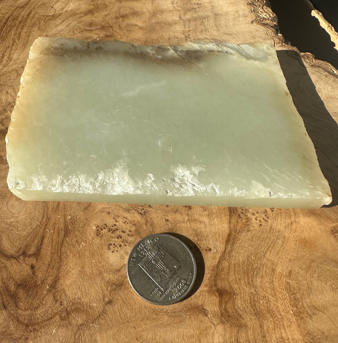 A rectangular pale green translucent stone slab with rough natural edges, photographed on a wood surface next to a coin for scale.