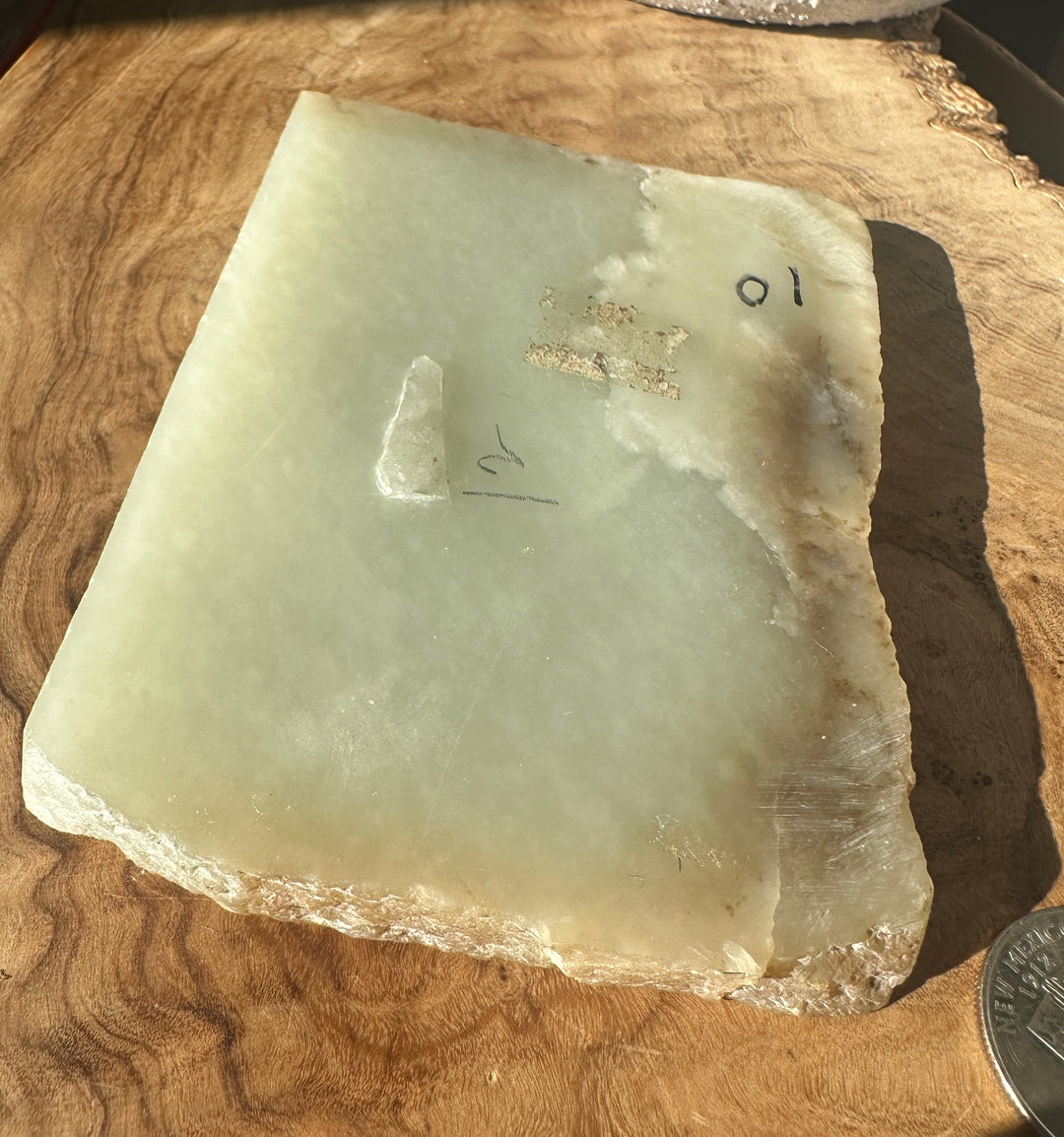 A raw, polished rectangular stone slab with a pale greenish-gray hue and rough, uneven edges, placed on a wooden surface with handwritten markings and a coin for scale.
