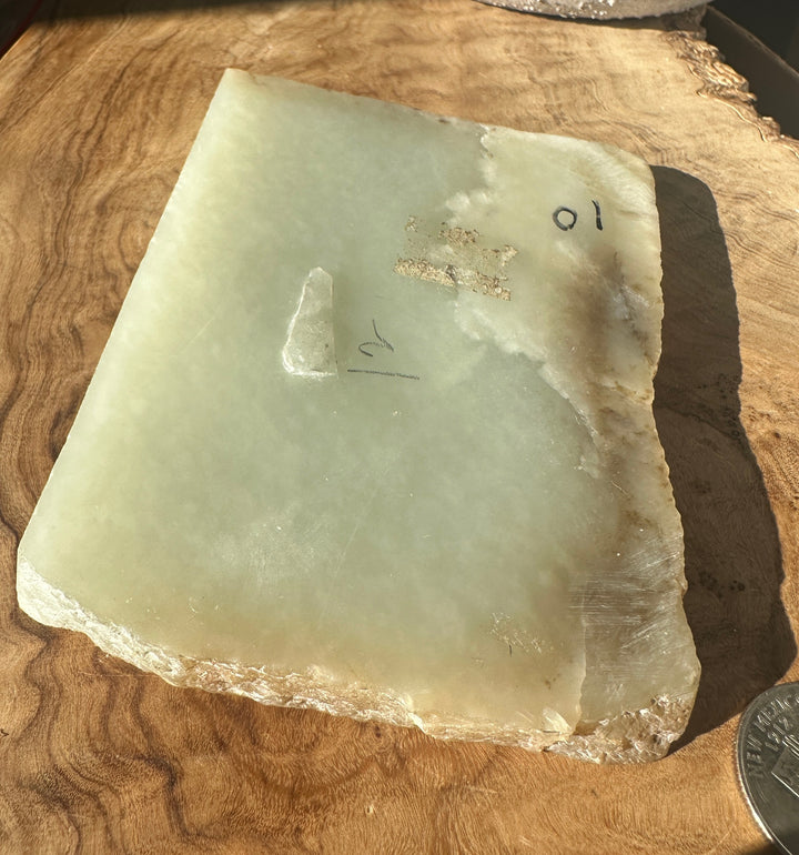 A raw, polished rectangular stone slab with a pale greenish-gray hue and rough, uneven edges, placed on a wooden surface with handwritten markings and a coin for scale.