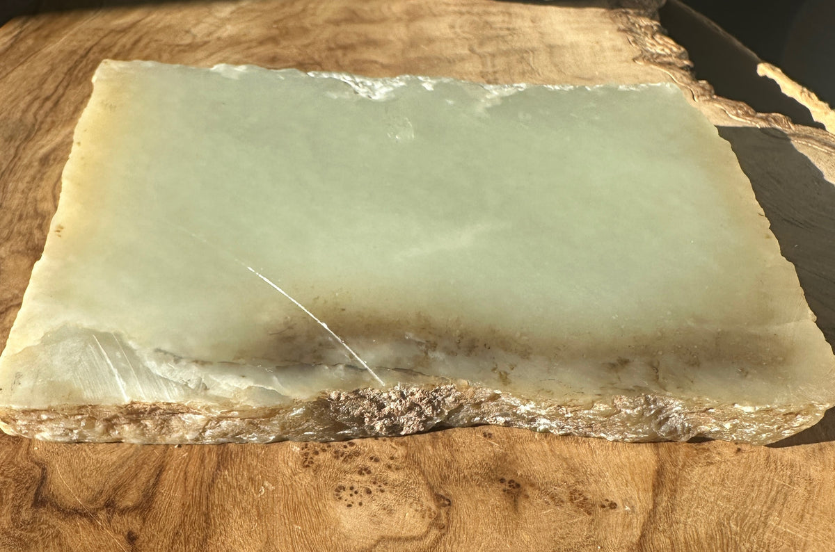 A rectangular jade slab with a pale green top surface and a rough, natural brown edge, displayed on a wooden surface.