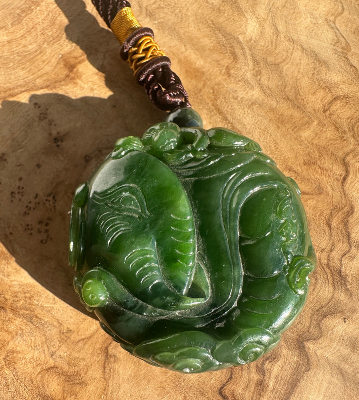 Green nephrite jade hand held pendant carved with elephant and monkey figures on braided brown and yellow cord against a tan background.