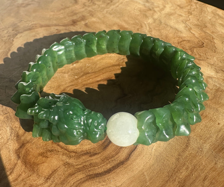 A carved green jade bracelet with a textured, segmented design and a smooth white central bead, displayed on a wooden surface.