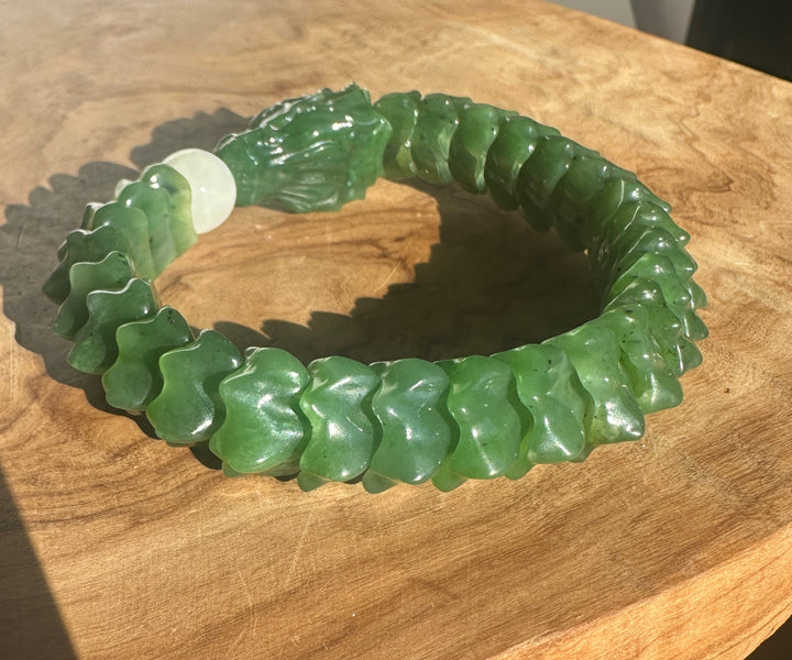 A carved green jade bracelet with a textured, segmented design and a smooth white central bead, displayed on a wooden surface.