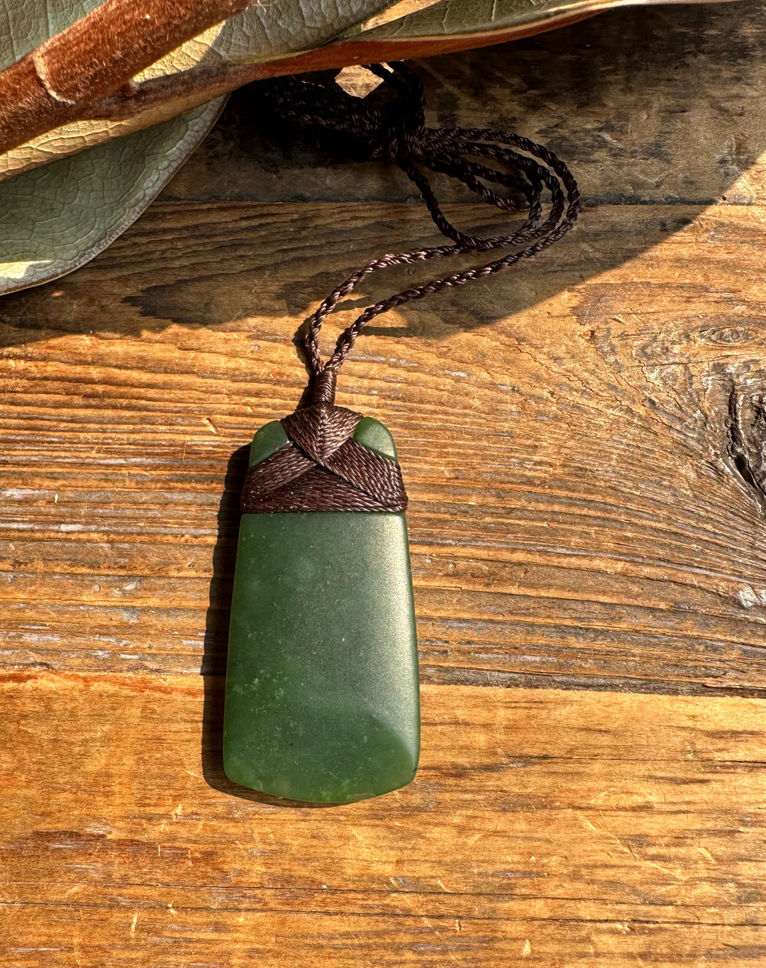 Dark green jade adze pendant with braided brown cord rests on a rustic wooden surface