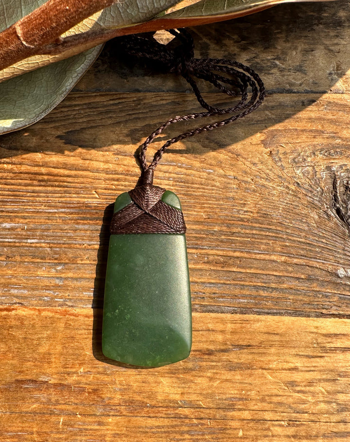 Dark green jade adze pendant with braided brown cord rests on a rustic wooden surface