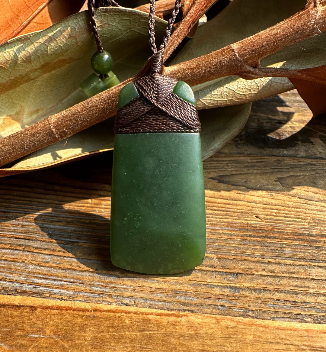 Dark green jade adze pendant with braided cord wrapping and green beads resting on wood with leaves
