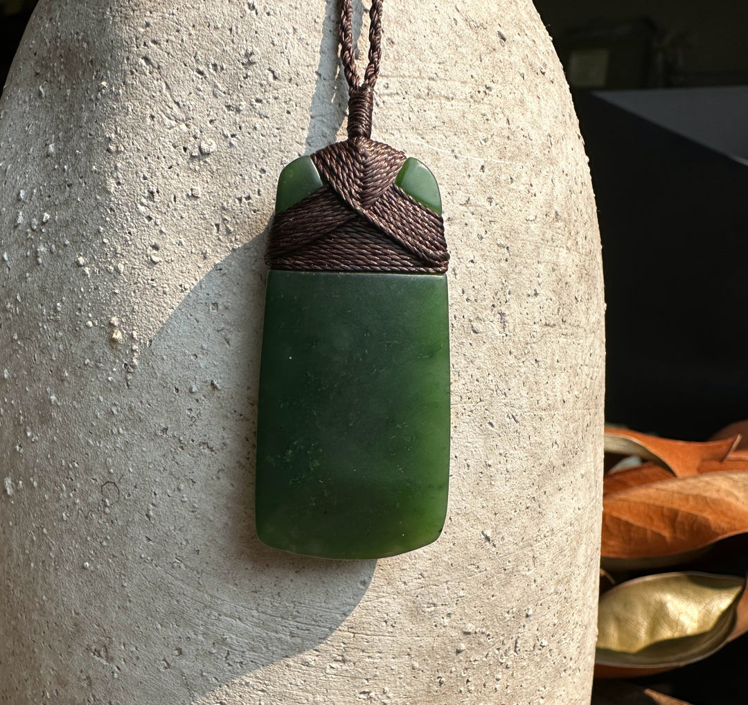 Dark green jade adze pendant wrapped in brown cord hangs against a light textured stone surface