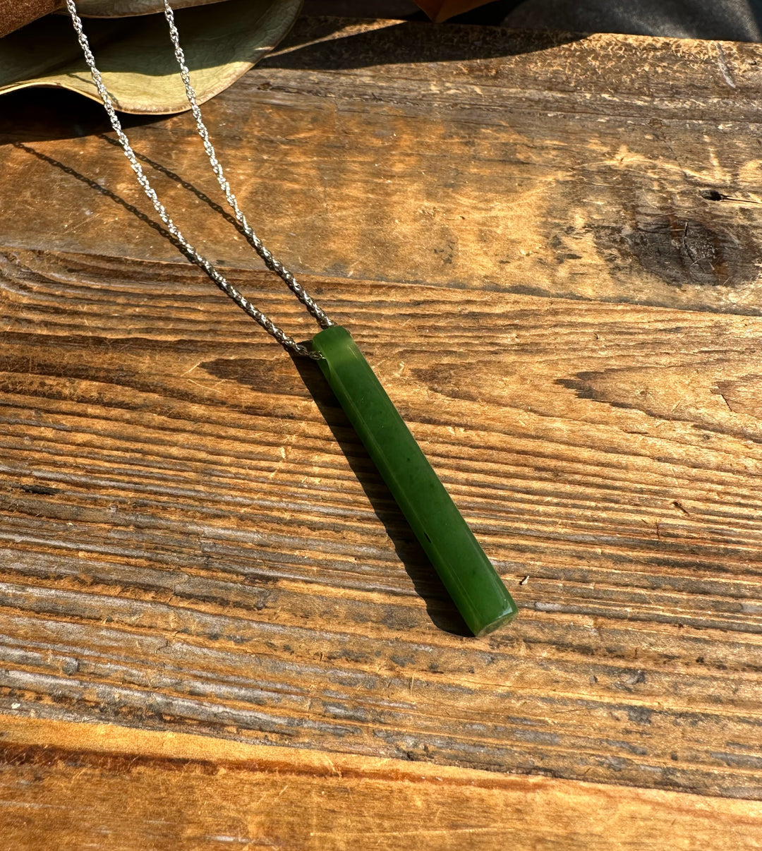 Thin green jade pendant on a silver chain resting on sunlit wood plank