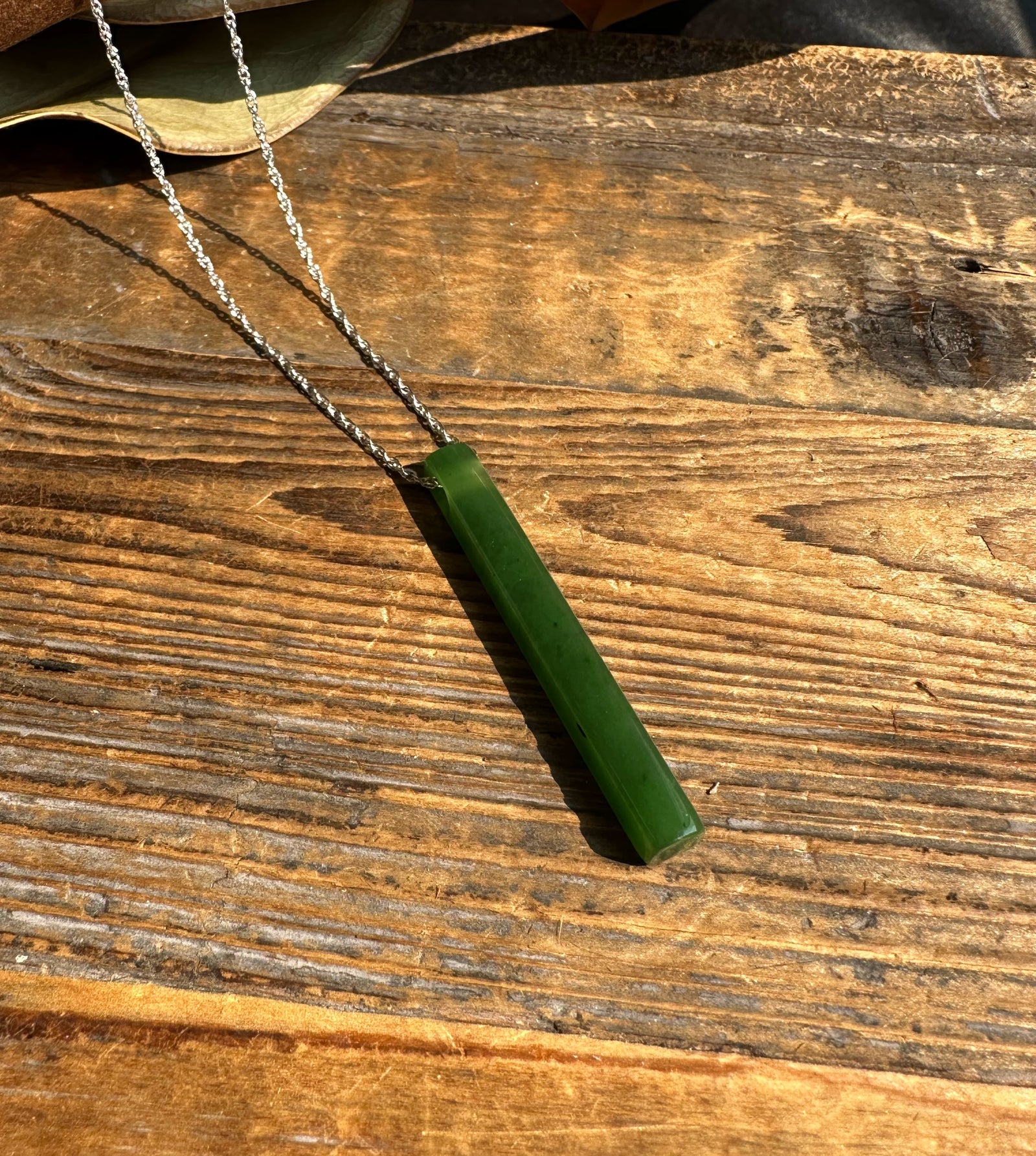Thin green jade pendant on a silver chain resting on sunlit wood plank