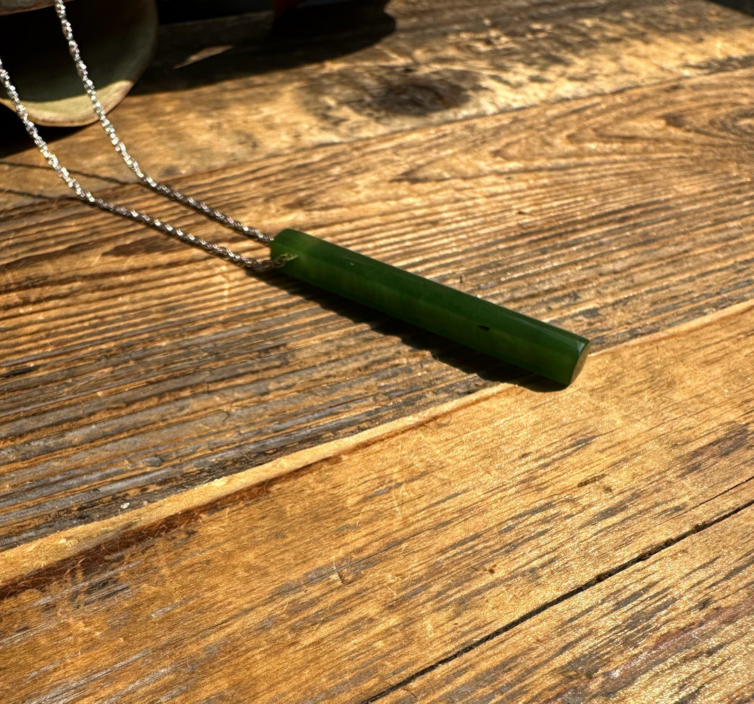 Slim green jade drop pendant resting on a rustic wooden surface with a twisted silver chain