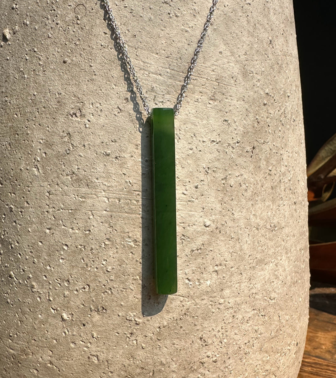 Green jade drop pendant with a round bead above on a silver chain against a textured stone surface