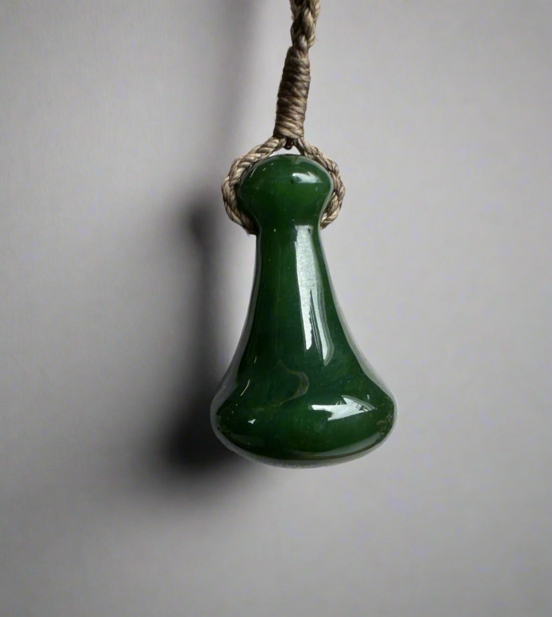 Close up of a green jade Poi Pounder pendant hung from braided twine against a neutral background.