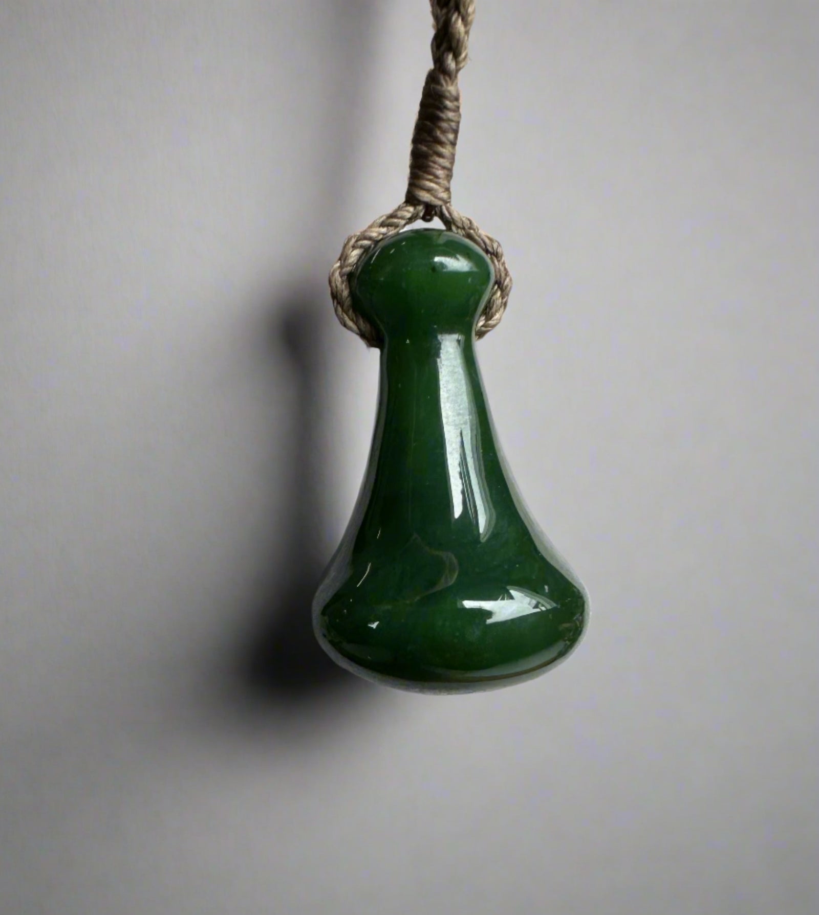 Close up of a green jade Poi Pounder pendant hung from braided twine against a neutral background.