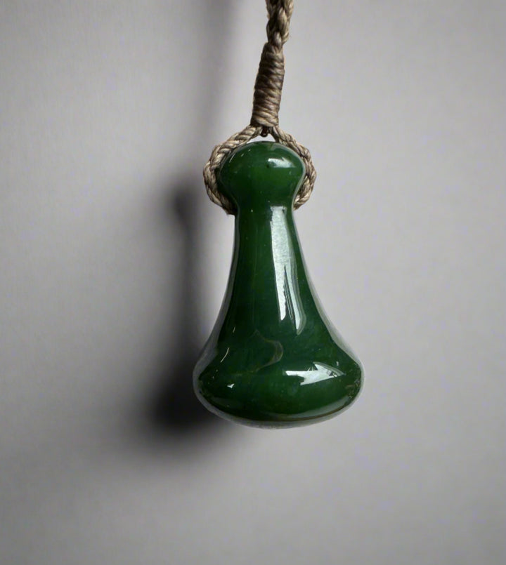 Close up of a green jade Poi Pounder pendant hung from braided twine against a neutral background.