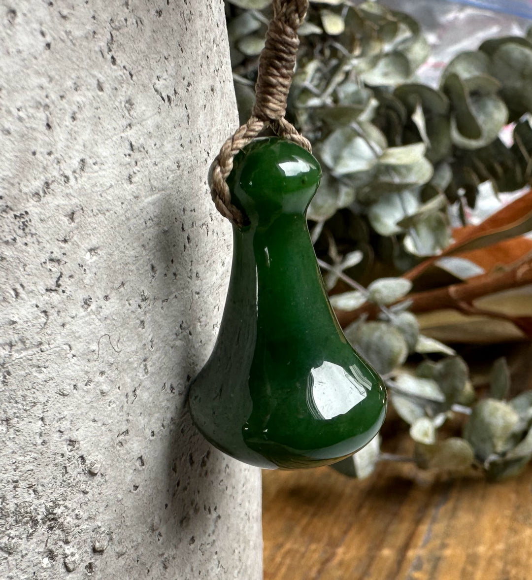 Poi Pounder Hawaiian Pendant in deep green stone hanging from a braided brown cord beside a textured tree trunk