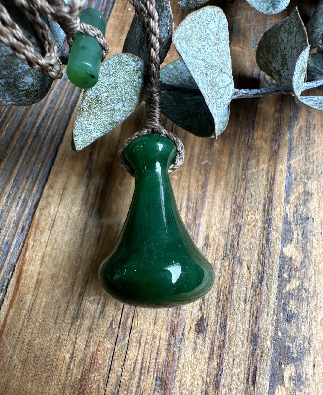 Close up of a deep green teardrop shaped Poi Pounder pendant on a knotted brown cord against a rustic wooden surface