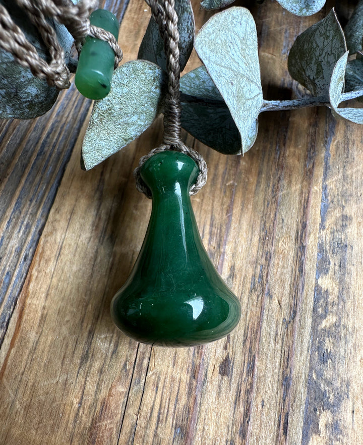 Close up of a deep green teardrop shaped Poi Pounder pendant on a knotted brown cord against a rustic wooden surface