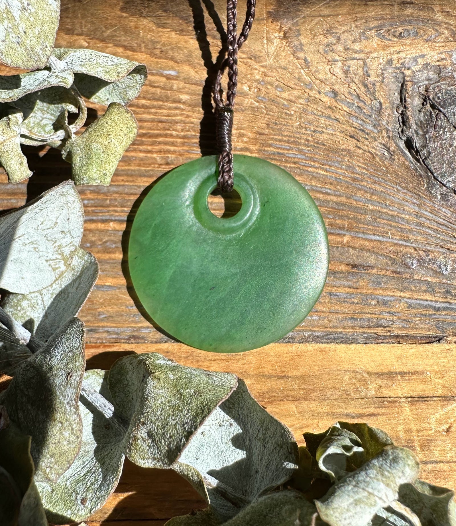 Jade round pendant 30mm with a central hole on a braided brown cord against a rustic wooden background.