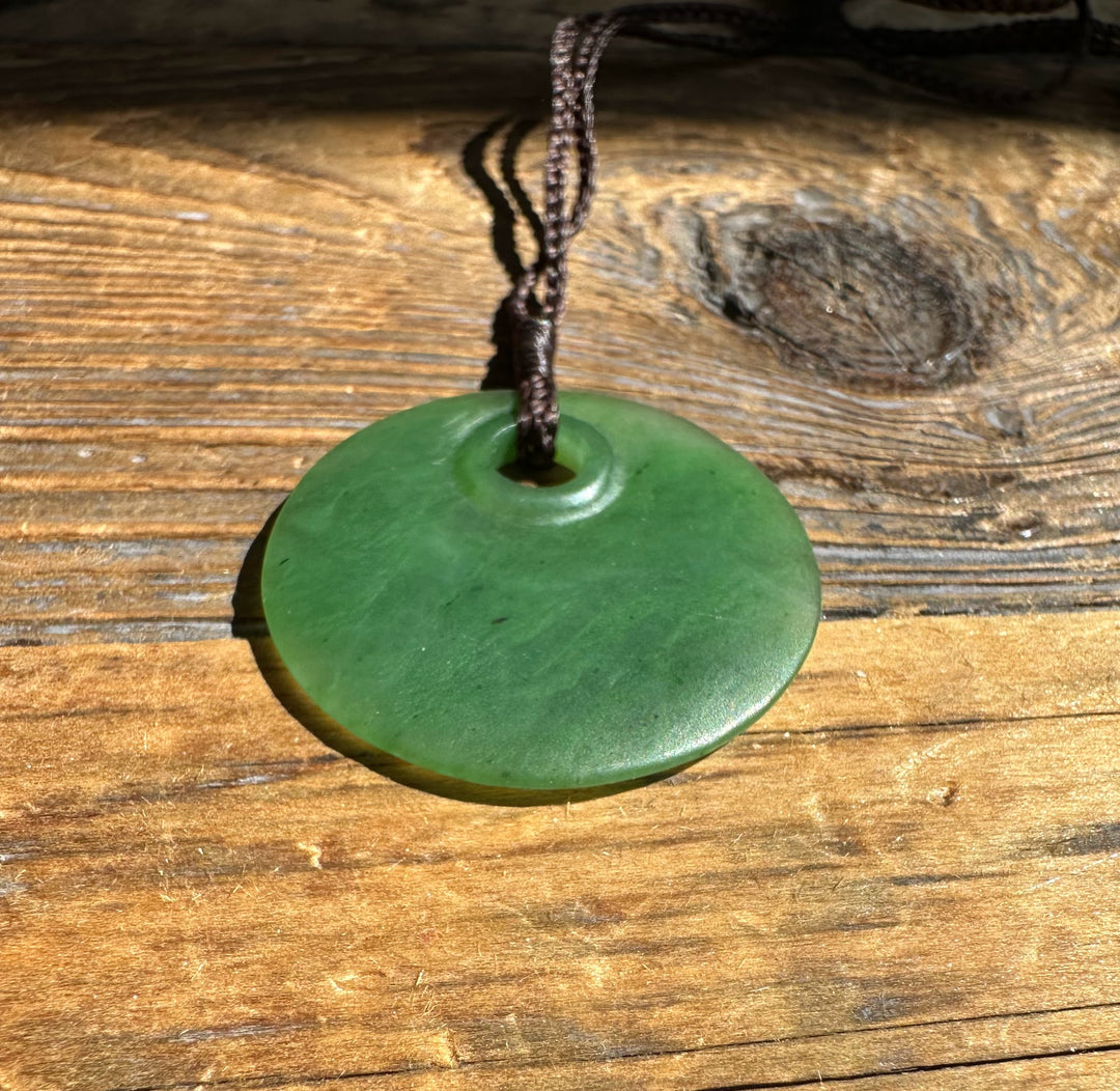 Jade round pendant 30mm with hole on a braided cord resting on sunlit wood.