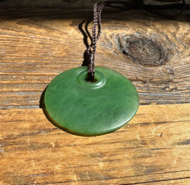 Jade round pendant 30mm with hole on a braided cord resting on sunlit wood.