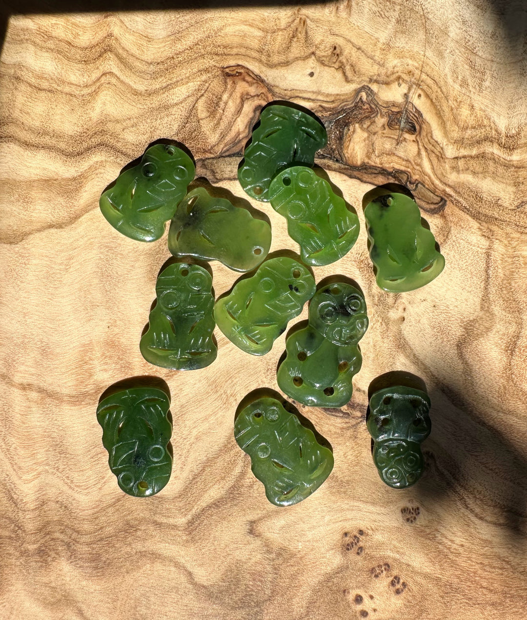 Eleven green jade tiki pendants arranged on wood with carved faces and drilled holes