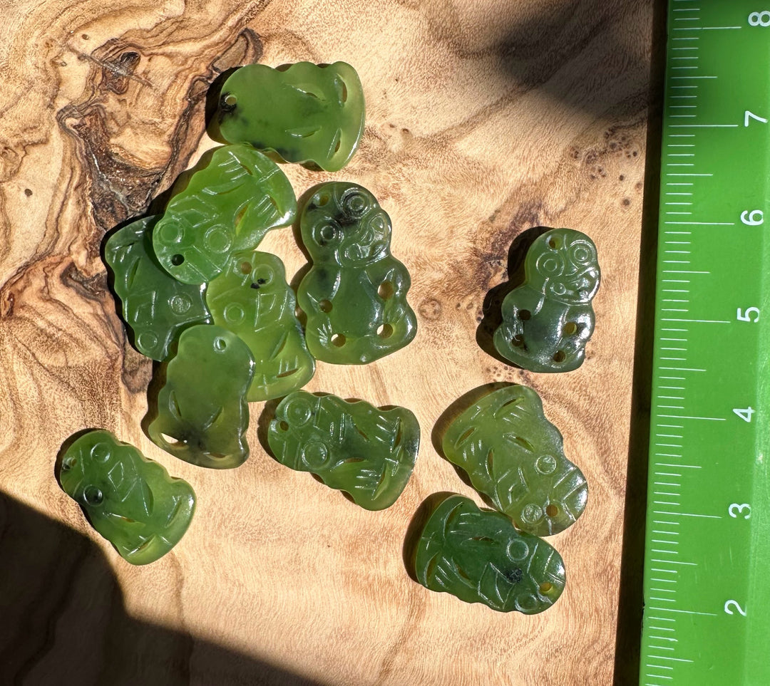 Green jade tiki pendants laid out on wood beside a measuring ruler for scale
