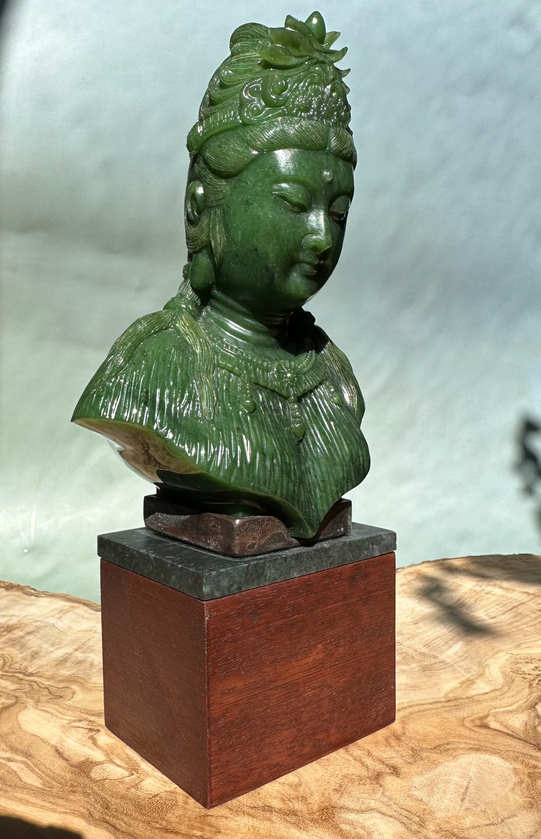 Green Guanyin statue viewed from the side, detailed robe texture and ornate crown on a dark slate top and wooden pedestal.