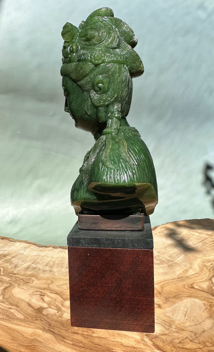 Side profile of a green Guanyin bust with an ornate crown and flowing robes on a dark stone plinth.