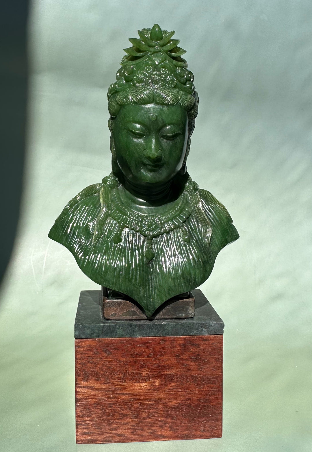 Front view of a green Guanyin bust with an elaborate crown and jewelry on a wooden base.