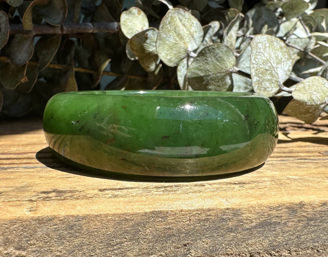 Glossy green Canadian Jade Wide Bangle resting on a sunlit wooden surface with a foliage backdrop