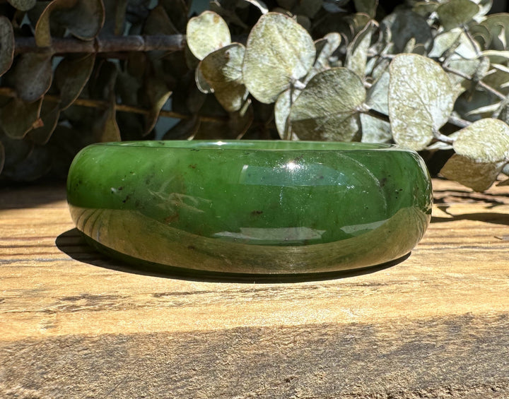 Glossy green Canadian Jade Wide Bangle resting on a sunlit wooden surface with a foliage backdrop
