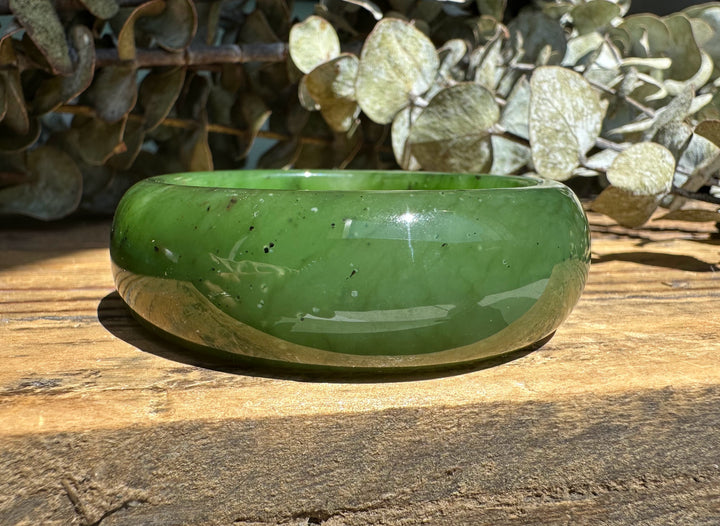 Emerald green jade wide bangle viewed from the side on a weathered wooden surface with leafy background