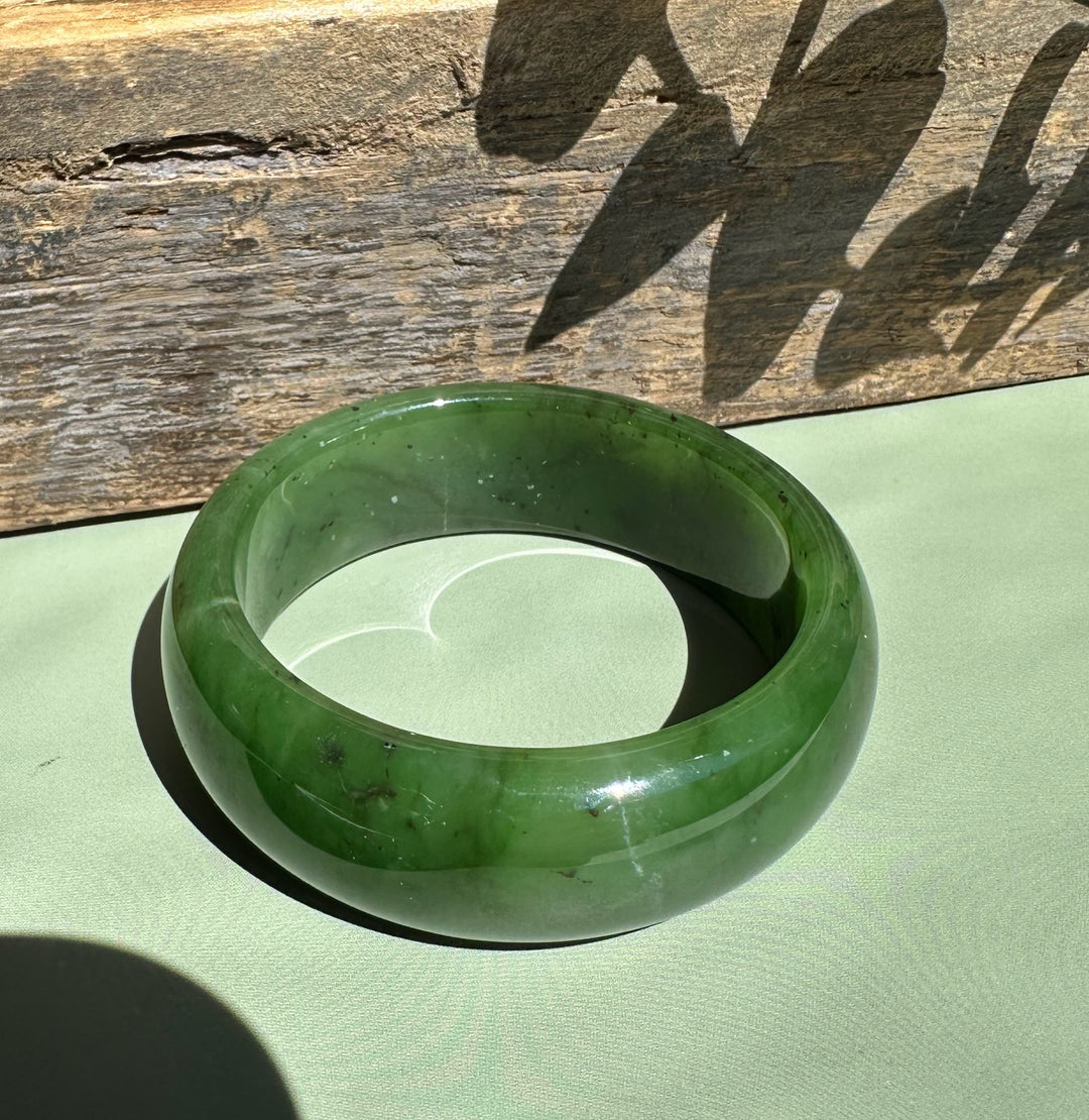 Canadian Jade Wide Bangle in deep green rests on a light green surface with a weathered wooden backdrop