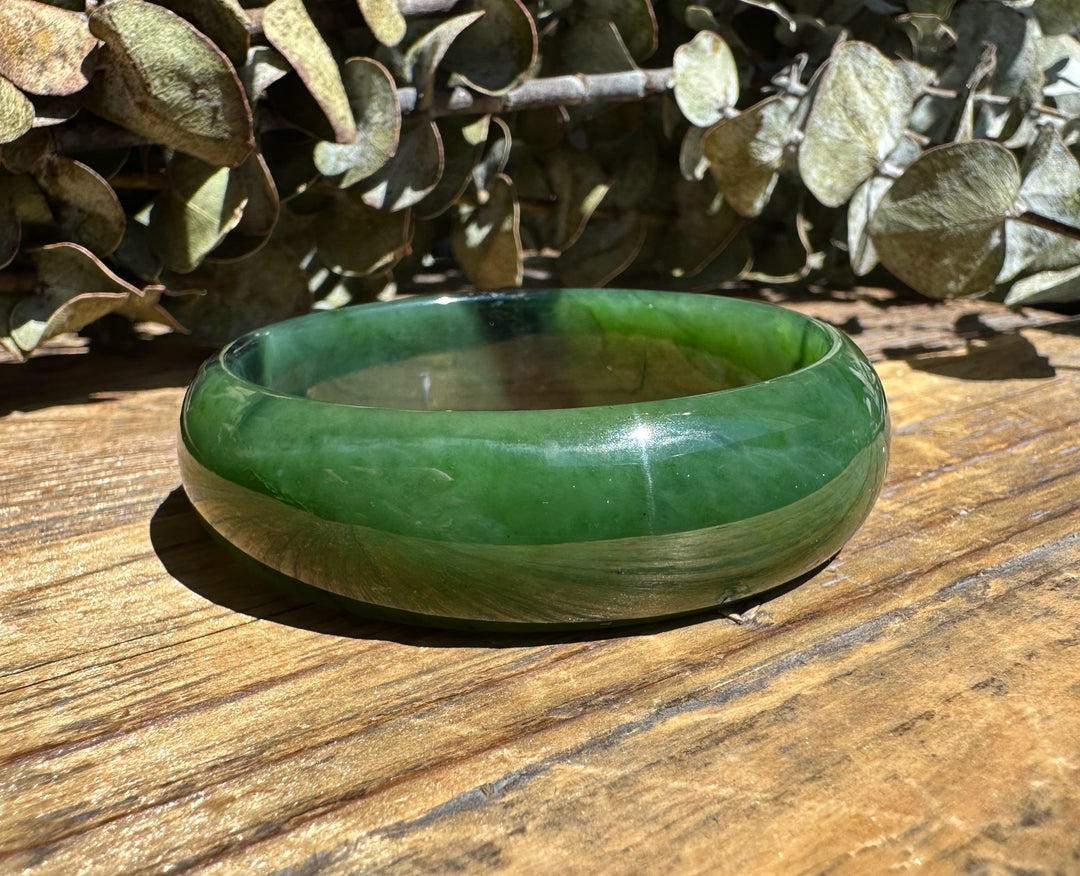 A Grade Canadian nephrite jade bangle with a chatoyant band on sunlit wood with dried eucalyptus behind