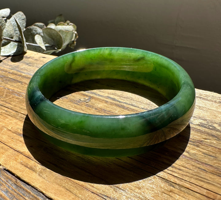 A Grade Canadian nephrite jade bangle with vibrant greens and banding resting on sunlit wood