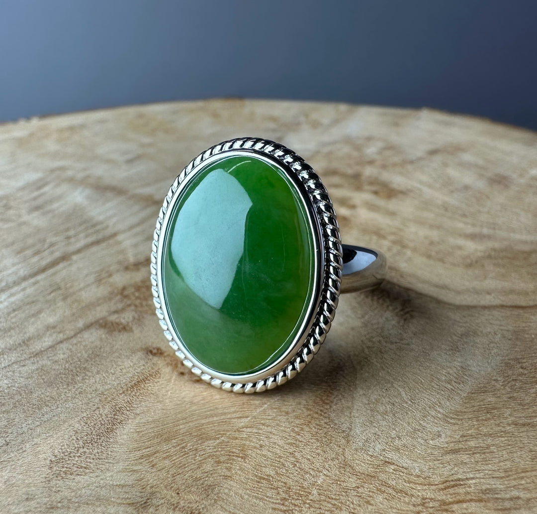 Green jade oval cabochon set in a silver ring with a rope edge on a wooden surface.