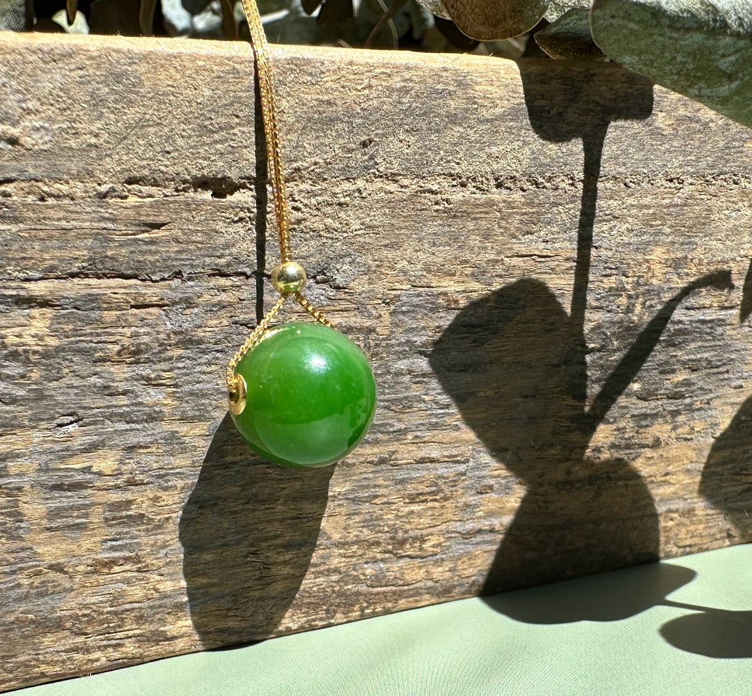 A bright green jade bead pendant with gold bail on a delicate chain against weathered wood