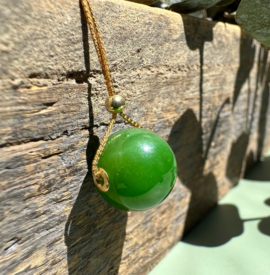 A+ Canadian Jade Bead Pendant 12mm with a gold bail on a slender golden chain against a rustic stone backdrop