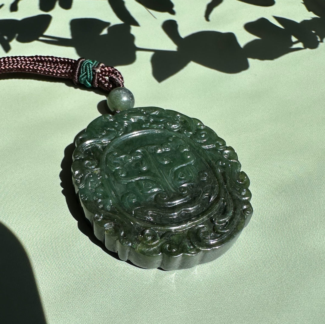 Round dark green jade Ku Lou pendant with ornate swirl border hanging from a braided brown cord