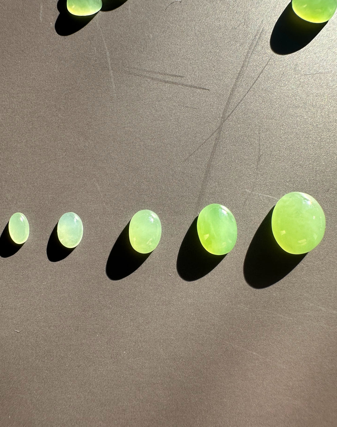 Line of apple green oval cabochons on a dark surface with strong shadows.
