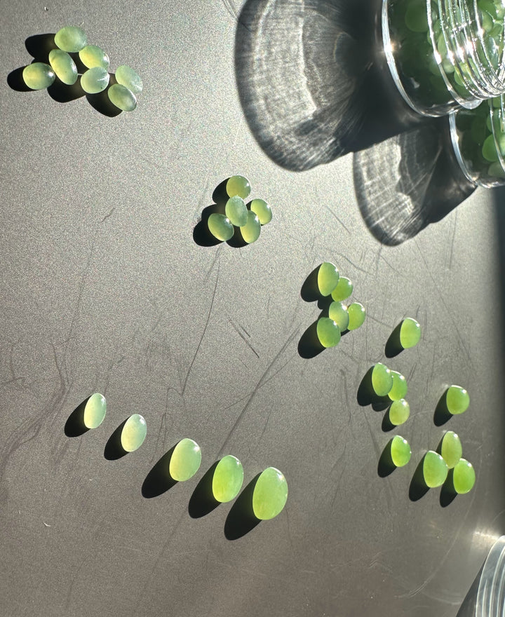 Assorted apple green oval cabochons in varying sizes scattered on a dark tabletop near a glass jar.