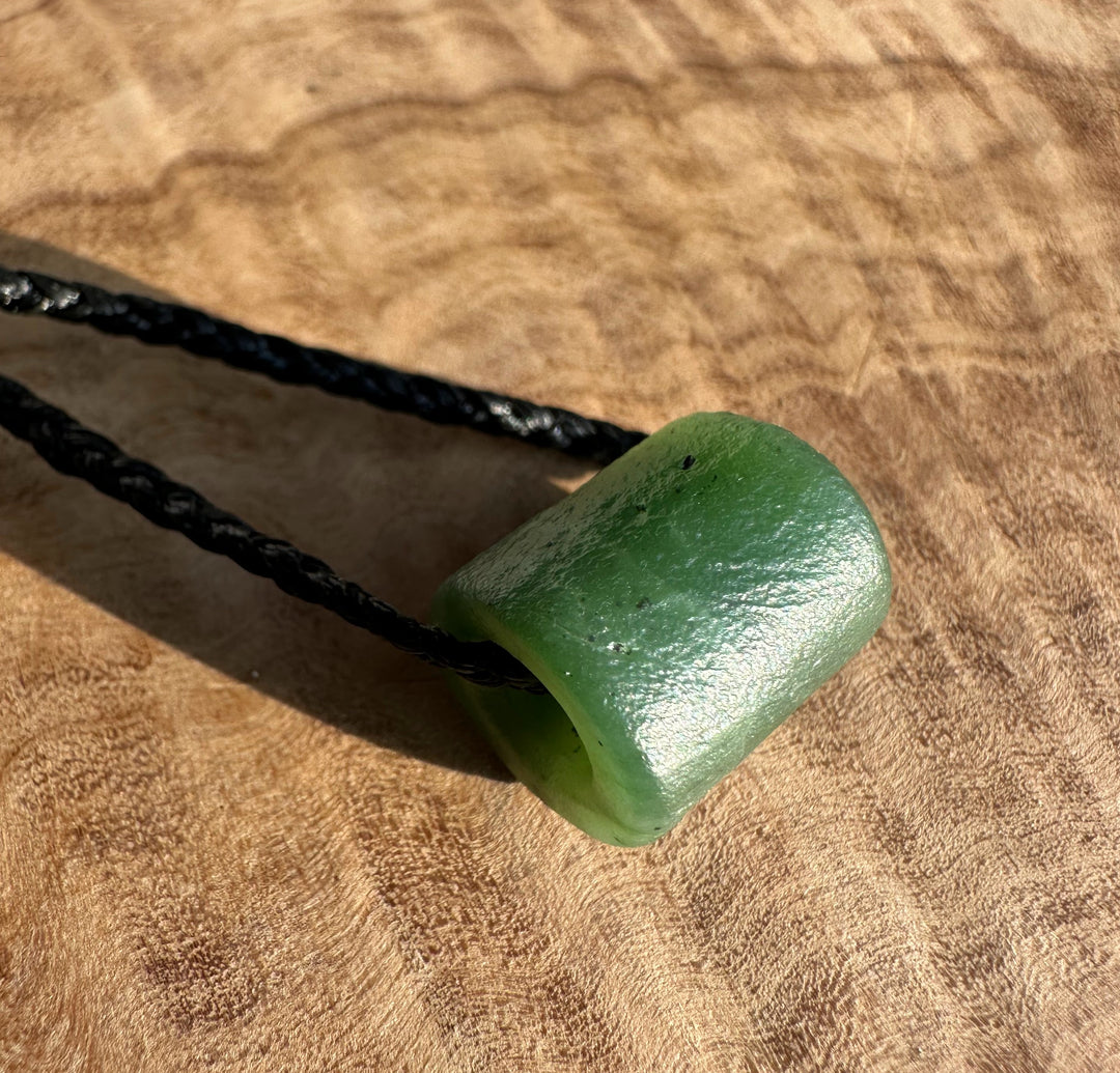 Side view of textured green jade pendant with a black braided cord looped through its hole on a wooden surface