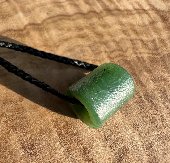Side view of textured green jade pendant with a black braided cord looped through its hole on a wooden surface